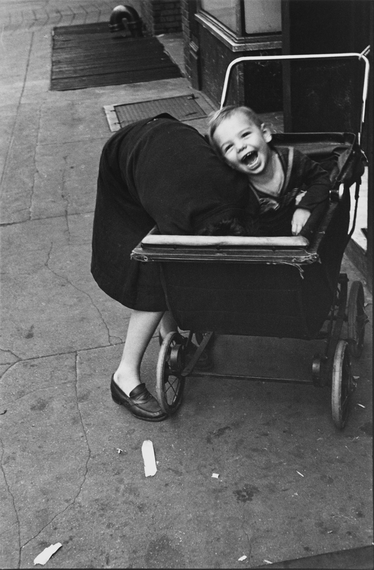Exposición Helen Levitt Fotografía en blanco y negro de un niño alegre riendo mientras juega en la calle. El niño está sentado en un cochecito, con un adulto parcialmente visible al fondo, inclinado sobre el cochecito. La imagen captura la espontaneidad y la naturaleza despreocupada de la infancia, típica de la fotografía callejera de Helen Levitt, centrada en escenas urbanas de Nueva York. La risa contagiosa del niño y el momento íntimo y sincero evocan una sensación de inocencia y energía juguetona, lo que encarna la capacidad de Levitt para transmitir emociones y experiencias humanas universales a través de momentos cotidianos.
